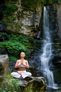 Beautiful young woman meditating in lotus position while doing yoga in the nature near waterfall